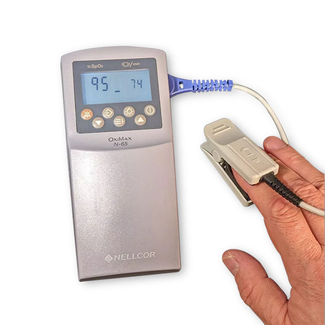 A hand holding a Covidien handheld pulse oximeter with a digital display, showing oxygen saturation levels, and a finger clip sensor attached to the finger.